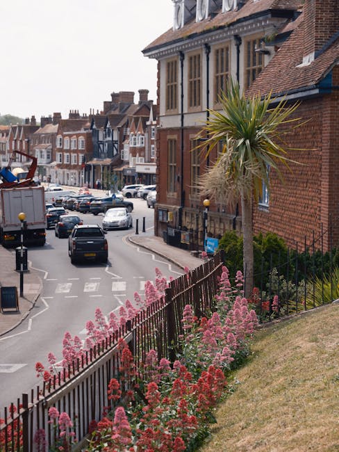 A street view in Loughton IG10 featuring a lush, landscaped area with vibrant pink and red flowers along a black metal fence, adjacent to a tall, slender palm tree with spiky green and brown fronds. I