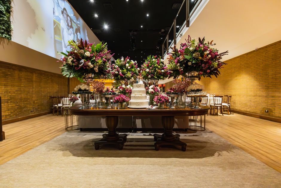 An elegant floral arrangement displayed on a dark wooden table in a spacious event hall with warm brick walls and light wooden flooring. The arrangement features large bouquets of pink, purple, and wh