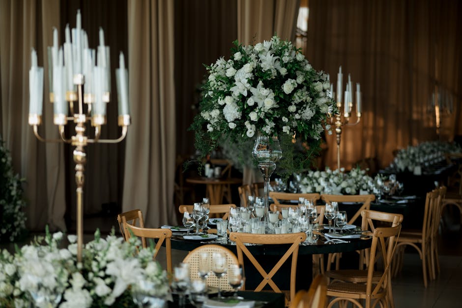 An elegant event table setup featuring a tall, lush floral centerpiece with a mix of white roses, lilies, and other delicate white flowers, accented by green foliage, placed in a clear glass vase. The