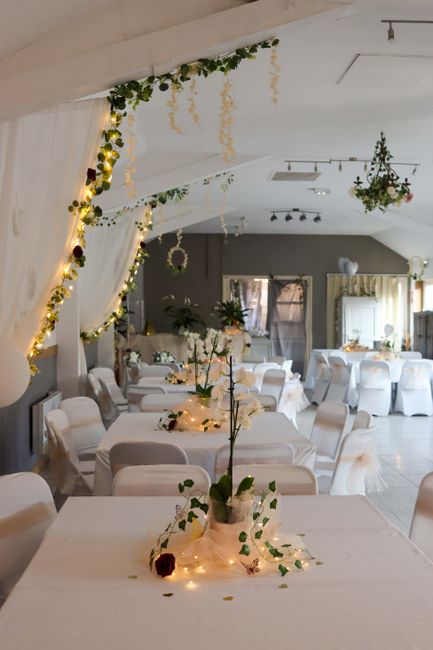 A wedding reception venue decorated with hanging greenery and string lights along a white draped ceiling. Tables are covered with white cloths and set with simple floral arrangements featuring white f