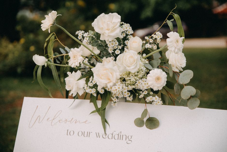 A floral arrangement featuring white roses, daisy-like chamomile flowers, and delicate white blooms, accented with soft eucalyptus leaves and wispy greenery. The bouquet is arranged in a loose, natura