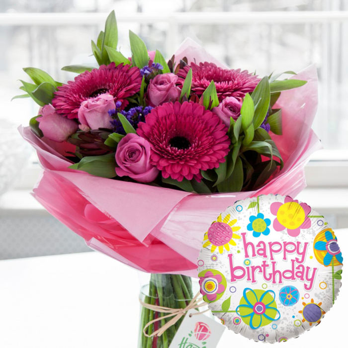 A joyful birthday flower and balloon duo is captured in crisp detail, as if freshly delivered to a home just off Loughton High Road. Dominating the scene is a generous bouquet arranged in a straight-sided glass cylinder vase, the stems visible through the water, neatly aligned by a skilled florist's hand. The flowers form a lush, rounded mass of colour, built around striking pink gerbera daisies in rich magenta and deep fuchsia shades. Their wide, symmetrical petals fan out from almost black central discs, creating bold focal points that draw the eye. Intertwined with the gerberas are numerous pink roses, ranging from blush to soft candy tones. Some roses remain tightly budded, holding their fragrance close, while others are beginning to open, revealing layered petals that suggest both elegance and affection. Small clusters of dark purple filler flowers, likely statice, peek between the larger blooms, giving subtle depth and texture, while a generous amount of fresh green foliage - broad, glossy leaves and finer sprigs - frames the entire arrangement, evoking the greenery of nearby Epping Forest. The base of the bouquet is wrapped in a collar of pale pink, translucent cellophane that crinkles softly and catches the light, forming a delicate skirt around the stems. Around the vase neck, a rustic piece of natural twine is tied into a simple bow, holding a small, off-white rectangular gift tag. The tag features a refined rose motif and understated branding that hints at a premium supplier without overshadowing the flowers. To the right and slightly in front of the vase, a round Mylar Happy Birthday balloon adds a playful flourish. The balloon's white background, lightly speckled in grey, is decorated with bright, stylised flowers and circular shapes in lemon yellow, sky blue, lime green, vivid pink, and deep purple, surrounding the words happy birthday written in bold, curving pink script. The softly blurred indoor background suggests a bright, welcoming room in Loughton or nearby Debden, ensuring the bouquet and balloon remain the undisputed stars of this celebratory composition, ready to delight any birthday recipient.