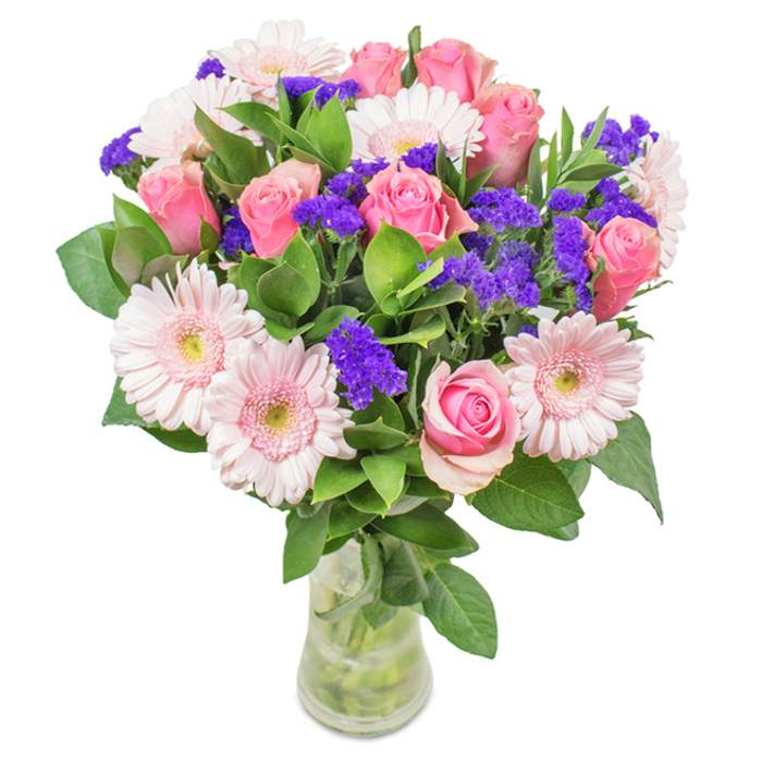 This vase arrangement was composed to feel like a brief walk through Loughton's quieter streets at dawn - soft, composed and full of discreet charm. The dominant signature is a cluster of pale pink Gerbera daisies, their broad, many-petalled heads and near-white centres creating a luminous halo of blush across the bouquet. Complementing them are several medium-pink roses, deliberately arranged at staggered heights; some remain in tight bud form while others open in classic spirals, their layered petals offering both visual rhythm and a gentle floral fragrance. Throughout the grouping, bright purple statice or limonium provides concentrated bursts of texture and colour, its papery clusters standing in lively contrast to the velvety, rounded blooms. Beneath and between the flowers, lush deep-green foliage creates volume and a verdant backdrop that feels fresh and cool, reminiscent of the leaves and mosses along nearby woodland paths. The stems are visible through a clear glass vase filled with water, the translucence and reflections adding a quiet, honest finishing touch to the composition. Thoughtfully balanced for scale and movement, this bouquet will sit well on a dining table or mantel in a Loughton home, or be carried as a modest but elegant gift for Mother's Day, anniversaries, or to mark a milestone. The arrangement aims to be both joyful and soothing: an everyday luxury that reads as crafted care, with colours and textures chosen to brighten a room without overwhelming its quiet atmosphere.